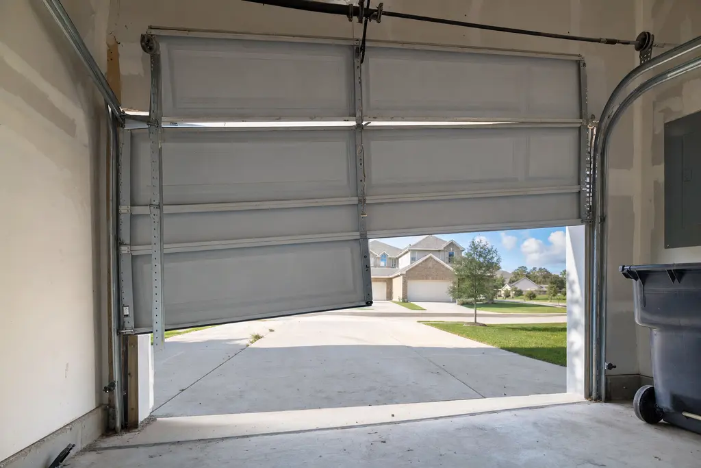 White steel garage door hanging crooked and partly open, one side out of its track, driveway visible beyond